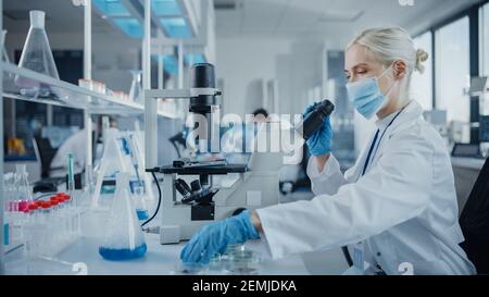 Modern Medical Research Laboratory: Portrait of Female Scientist Wearing Face Mask Using Microscope, Analysing Test Samples. Advanced Scientific Lab Stock Photo