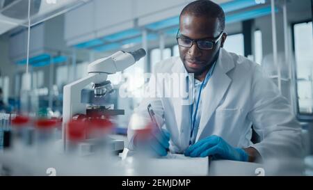 Modern Medical Research Laboratory: Portrait of Male Scientist Using Microscope, Writing Down Analysis Information. Advanced Scientific Lab for Stock Photo