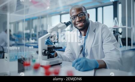 Modern Medical Research Laboratory: Portrait of Male Scientist Using Microscope, Charmingly Smiling on Camera. Advanced Scientific Lab for Medicine Stock Photo