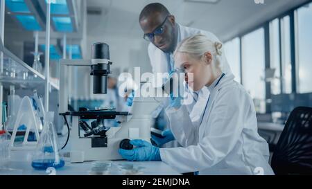 Modern Medical Research Laboratory: Two Scientists Working Together Using Microscope, Analysing Samples, Talking. Advanced Scientific Lab for Medicine Stock Photo