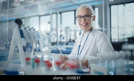 Medical Research Laboratory: Portrait of a Beautiful Female Scientist Writing Down Data, Smiling on Camera. Advanced Scientific Lab for Medicine Stock Photo