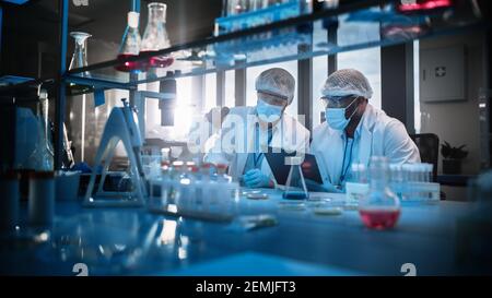 Modern Medical Research Laboratory: Two Scientists Wearing Face Masks use Microscope, Analyse Sample in Petri Dish, Discuss Innovative Technology Stock Photo