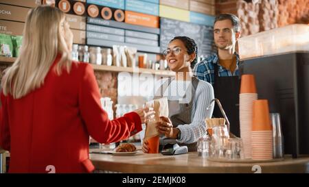 Joyful Multiethnic Diverse Woman Gives a Payment Terminal to Customer Using NFC Technology on Smartphone. Customer Uses Mobile to Pay for Take Away Stock Photo