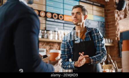 African American Customer Pays for Coffee and Pastry with Contactless NFC Payment Technology on Smartphone to a Handsome Barista in Blue Checkered Stock Photo