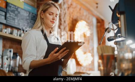 Beautiful Young Caucasian Coffee Shop Manager with Blond Hair is Making Notes on a Tablet Computer and Orders Inventory Items for the Menu in a Cozy Stock Photo