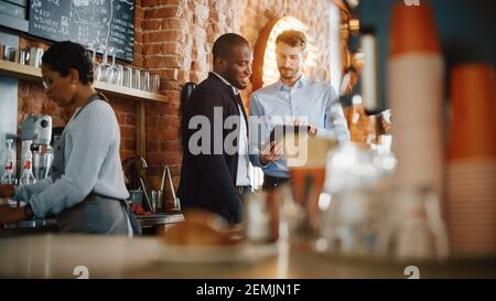 Multicultural Coffee Shop Owners Meeting Behind the Counter and Working on Tablet Computer and Checking Inventory in a Cozy Loft-Style Cafe Stock Photo