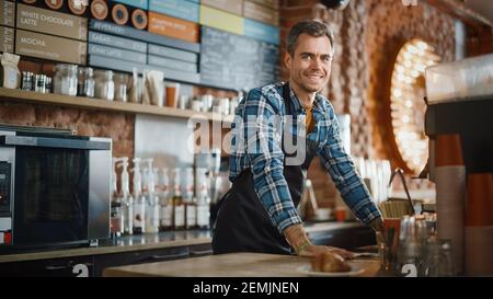 Handsome Barista in Checkered Shirt and Black Apron is Making a Cup of Cappuccino in Coffee Shop Bar and Smiling on Camera. Portrait of Happy Employee Stock Photo