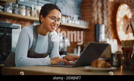 Young and Beautiful Latina Coffee Shop Owner is Working on Laptop Computer and Checking Inventory in a Cozy Cafe. Successful Restaurant Manager Stock Photo