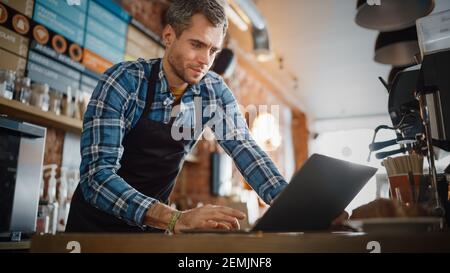 Young and Handsome Coffee Shop Owner is Working on Laptop Computer and Checking Inventory in a Cozy Cafe. Happy Restaurant Manager in Checkered Shirt Stock Photo