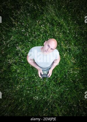 Man stood flying drone holding controller looking up to sky photo taken from above camera pointing down to him standing in long grass field on bright Stock Photo