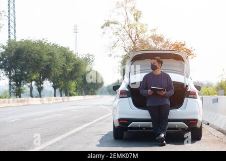 Asian traveler man wearing face mask and sitting on car with hightway road background Stock Photo