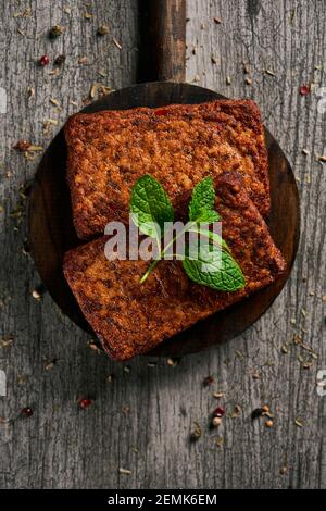 high angle view of some cooked tofu fillets in a round dark wooden tray, on a gray rustic wooden table sprinkled with different spices and herbs Stock Photo