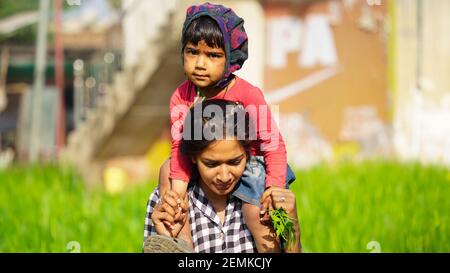 A little Indian baby sits on the shoulders of a smiling older sister. Cute baby girl playing in green field, winter season concept in village Stock Photo