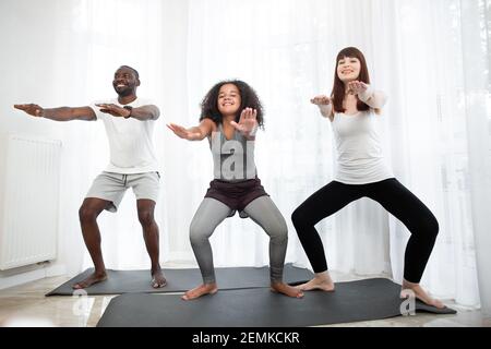 Active and healthy. Beautiful teenage mixed race girl and her parents in sports clothing doing squats and taking similar positions on the mat at home. Stock Photo