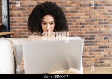 Portrait of a young African American female freelancer with Afro hairstyle sitting on the couch and working on project, watching movie on laptop, studying, blogging, resting and chatting online Stock Photo