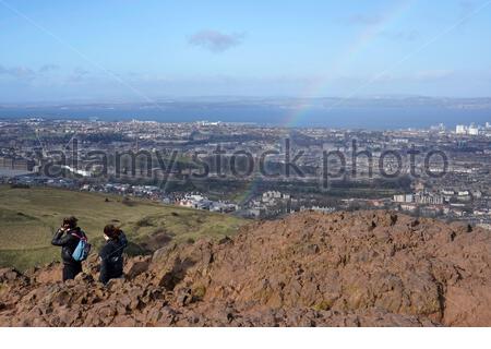 Edinburgh, Scotland, UK. 25th Feb 2021. People enjoying the sun and outdoors in Holyrood Park, on the summit of Arthurs Seat a rainbow appears after a rain shower.  Credit: Craig Brown/Alamy Live News Stock Photo