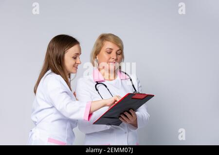 Quality medical services. Female doctors in uniform taking notes to clipboard while standing in hospital corridor. Medical conference on gray Stock Photo