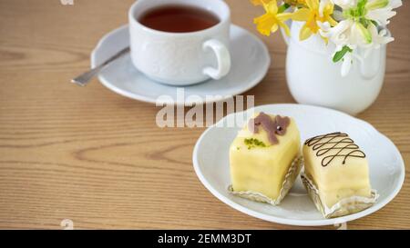 Cup of tea and Easter dessert- lemony yellow marzipan petit fours on white porcelain plate for  breakfast and daffodils on the wooden table. Close up, Stock Photo