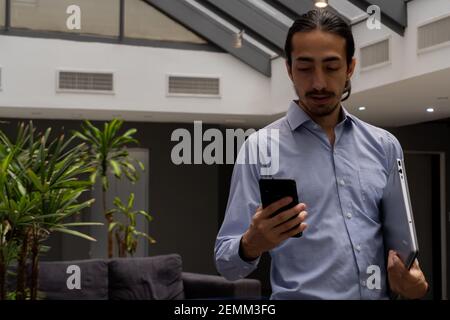 man in home office walking while checking his smart phone and holding his laptop in the other arm. life style Stock Photo
