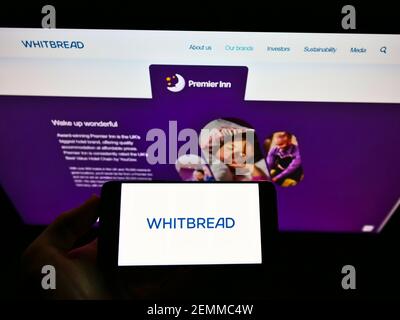 Person holding smartphone with logo of British hospitality company Whitbread plc on screen in front of website. Focus on phone display.