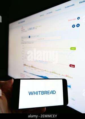 Person holding cellphone with logo of British hotel company Whitbread plc on screen in front of webpage with stock chart. Focus on phone display. Stock Photo