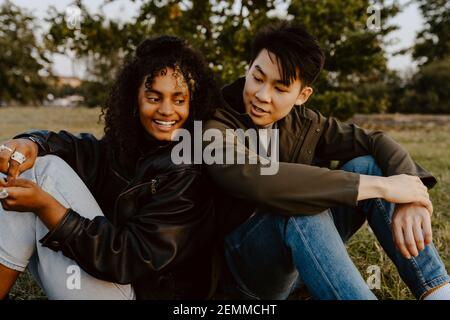 Happy friends sitting side by side in park during sunset Stock Photo