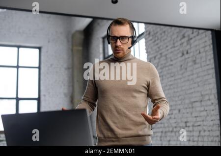 Portrait of young businessman in a headset looking on the laptop, hipster customer service representative standing in the modern office, actively thinking and concentrating on the customers problem Stock Photo