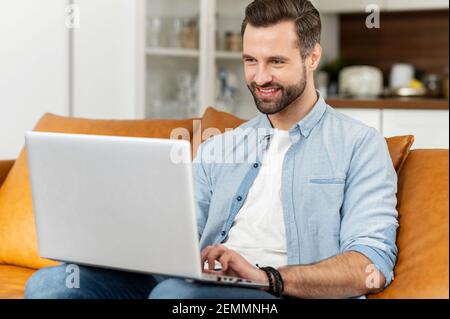 Smiling young bearded hipster man sitting on the couch, flirting, typing a message on the laptop, video-calling with friends or girlfriend online, chatting in social media network, using dating app Stock Photo