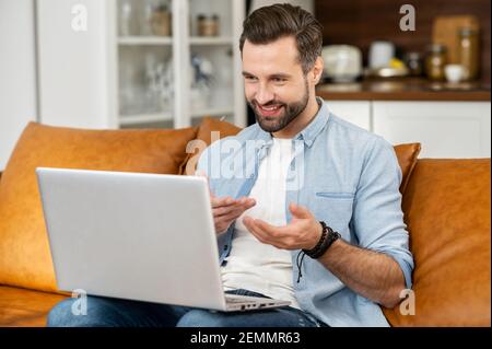 Smiling young bearded hipster guy tutor or a blogger sitting on the couch, streaming video online on the laptop, talking and explaining the strategy and gesturing, chatting in social media network Stock Photo