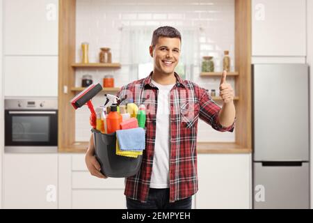 Smiling young man holding a bucket full of cleaning supplies and gesturing a thumb up sign in a modern kitchen Stock Photo