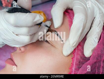 Beautician in white gloves making permanent make-up correction eyebrows shape. Female master using special needle, injects pigment into skin. Eyebrow Stock Photo