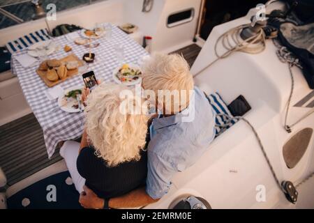High angle view of senior couple taking selfie on mobile phone in sailboat during sunset