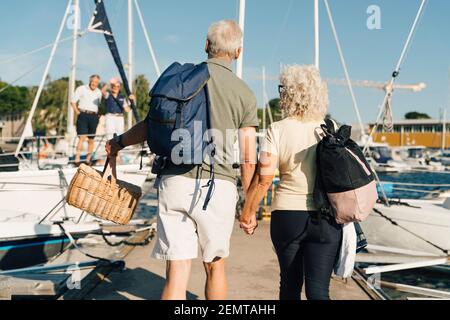 Rear view of senior couple holding hands walking on pier at harbor Stock Photo