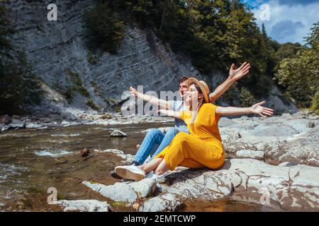 Couple of tourists relaxes by mountain river enjoying landscape. Travelers sit on rock with arms raised. Summer vacation trip Stock Photo