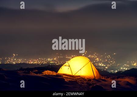 Yellow tent lighted from the inside against the backdrop of glowing city lights in fog. Amazing snowy landscape. Tourists camp in winter mountains. Travel concept Stock Photo