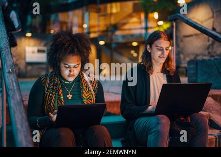 Young male and female students using laptop while sitting on steps in campus at night Stock Photo