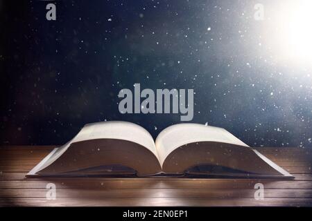 Open old book on wooden table and dark background with light shine and flying particles with mystical and imaginative look Stock Photo
