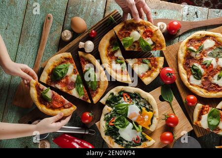 Slice of fresh pizzas child and mama hold with green basil, tomatoes, mushrooms and egg on old painted wood overhead view Stock Photo