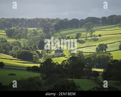 a view across landscape with patchwork of green farm fields, leafy trees and dry-stone walls below grey sky in rural hilly Cumbria, England, UK