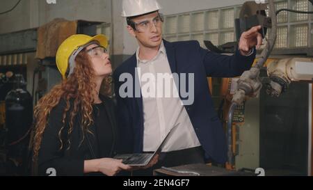 Caucasian maintenance engineer man and diverse female factory workers in hardhats talking and present or explain robot welding machine on plant, Manag Stock Photo