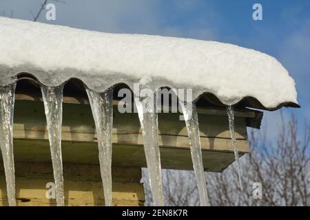 Large icicles, glittering in the sun in winter, are hanging from the roof are like roof decorations. Beautiful icicles along the roof. Stock Photo