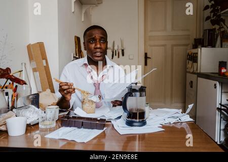 Thoughtful male entrepreneur with paper document having noodles at table in living room Stock Photo