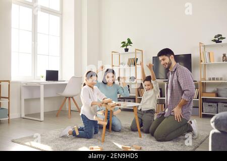 Mom, dad, daughter and son play game sitting at a table from which a wooden tower falls. Stock Photo