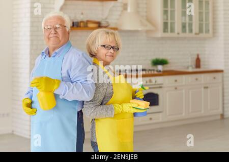 Ederly couple holding cleaning detergents and standing with their backs to each other in the kitchen Stock Photo