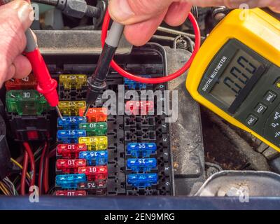 Man’s hands holding red and black probes from a digital multimeter on a 15 amp fuse, among many in a vehicle fuse box, to test an electrical circuit. Stock Photo