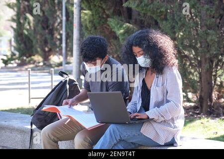 Two latin students wearing protective face mask studying together sitting on a bench outdoor. New normal at university campus. Stock Photo