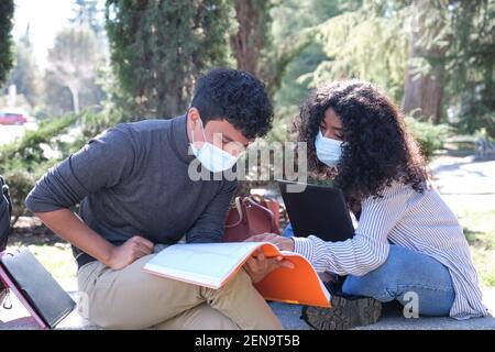 Two latin students wearing protective face mask studying together sitting on a bench outdoor. New normal at university campus. Stock Photo