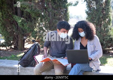 Two latin students wearing protective face mask studying together sitting on a bench outdoor. New normal at university campus. Stock Photo