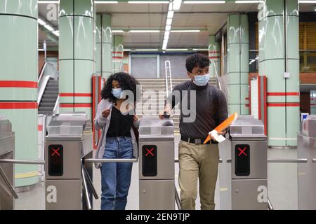 Young latin couple wearing protective face mask walking through subway turnstile at the train or metro station. New normal at public transport. Stock Photo