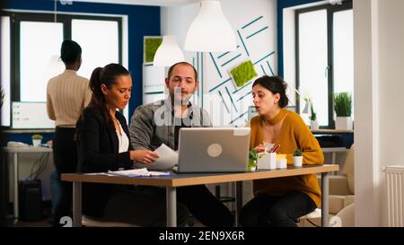 Enthusiastic concentrated business colleagues discussing holding documents and charts, analyzing company financial reports using laptop while multicultural team working together in modern office. Stock Photo
